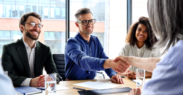 A professional man and woman shake hands at a meeting table in a brightly lit office space. Beside them, two colleagues look on, smiling. Glasses of water and business documents sit on the table.